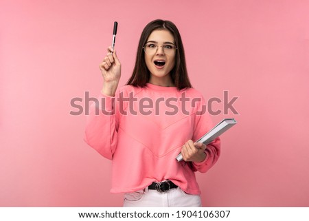 Photo of attractive woman in glasses holds book and pencil, looks quick-witted. Student got idea. Wear casual pink t-shirt white pants isolated pink color background