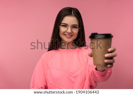 Photo of attractive woman in glasses holds coffee and takes selfie. Student wears casual pink t-shirt white pants isolated pink color background