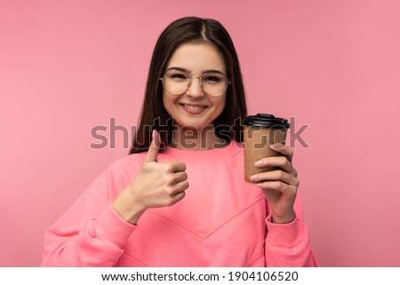 Photo of attractive woman in glasses holds coffee and gives thumb up. Wears casual pink t-shirt white pants isolated pink color background