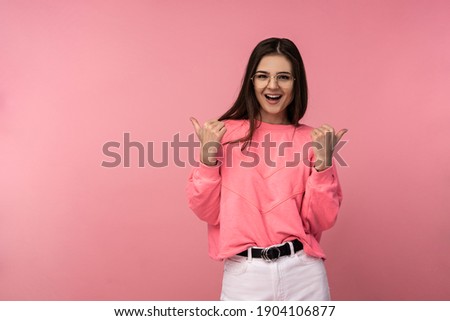 Photo of attractive young woman in glasses smiles and gives thumbs up. Wears casual pink t-shirt white pants isolated pink color background