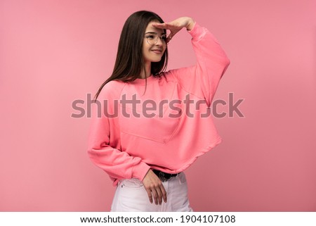 Photo of attractive young woman in glasses surprised, look into the distance. Wears casual pink t-shirt white pants isolated pink color background