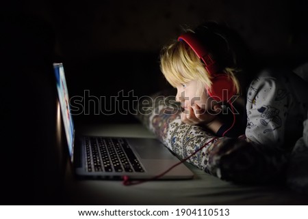 Cute preschool boy wearing headphones is watching cartoon movie by laptop before sleeping in bed or early in the morning right after waking up. Overuse and addiction kids from gadgets. 