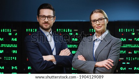 Portrait of two smart and successful programmers, man and woman, standing on background of running servers looking at camera with arms crossed.