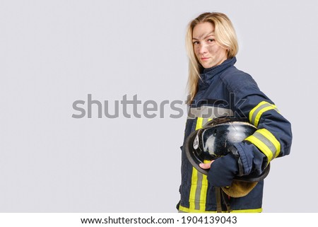 Young brave woman with dirty face in uniform of firefighter holds hardhat in hand and looking at camera isolated on gray background. Copy space for text 