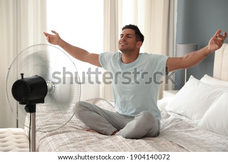 Man enjoying air flow from fan on bed in room. Summer heat Royalty-Free Stock Photo #1904140072
