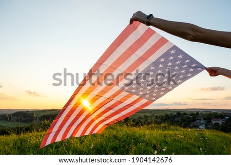 Human hands holding waving american national flag in field at sunset.