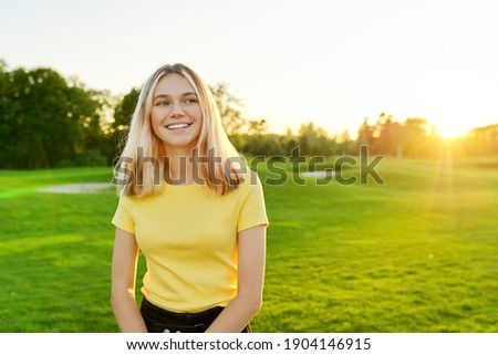 Outdoor portrait of smiling teenage girl 16, 17 years old in yellow T-shirt, on green sunny lawn. Sunset, grass background for copy space