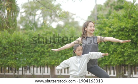 Happy asian mother and daughter doing yoga exercise on mat in grass park at the day time. People having fun outdoors. Concept of friendly family and of summer vacation. Healthy lifestyle.