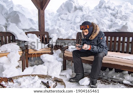 Teenager dressed in ski or snowboard fashion mask goggles uses a smartphone. Mountain landscape. Extreme adventure. Winter ski resort