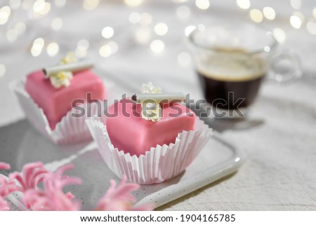 Valentine petit fours with marzipan icing and cream flowers. Espresso coffee in glass cup. Happy Valentine's day Garland of lights on ivory, off white textile tablecloth. Pink hyacinth flower.