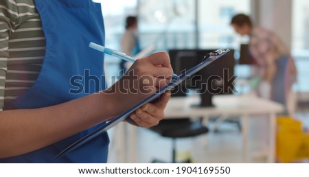 Team of professional janitors in uniform cleaning office. Close up of woman cleaner checking list on clipboard supervising cleaning service team work in modern office Royalty-Free Stock Photo #1904169550