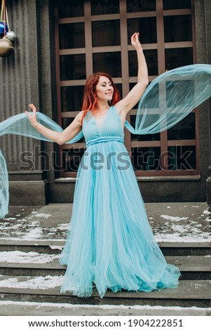 
beautiful bride with red hair in a blue long dress on the background of the city. valentine's day, wedding, winter, princess