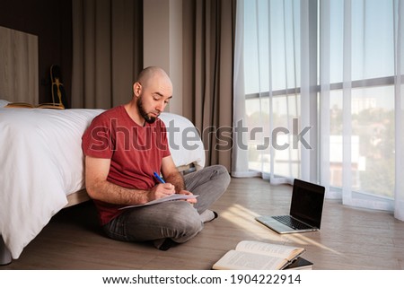 An adult bald man sits cross-legged on the floor and learns using a laptop. Indoor. The concept of online courses and online learning