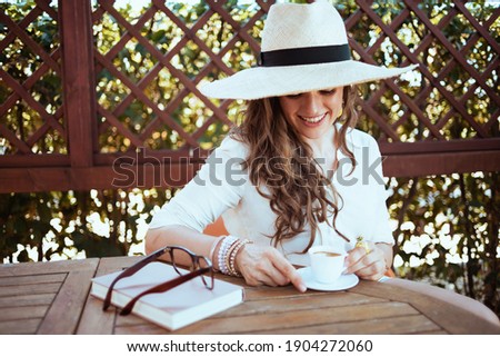 happy trendy middle aged woman in white shirt with book, hat and eyeglasses sitting at the table drinking coffee in the patio.