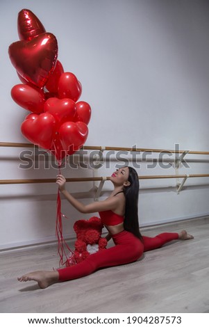 A young woman trainer in a red sports uniform does stretching in a sports hall near her there are heart-shaped balloons