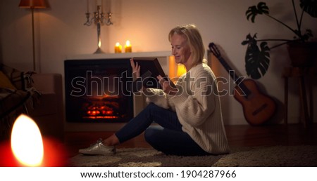 Happy mature woman looking at photograph in frame at home. Portrait of smiling senior lady sitting on carpet near fireplace and holding framed family photo