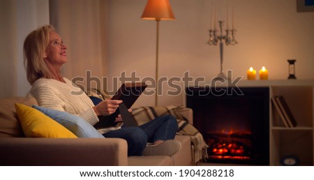 Smiling senior woman sitting o couch looking at photo in frame. Side view of cheerful aged lady relaxing on sofa in living room holding framed family photograph