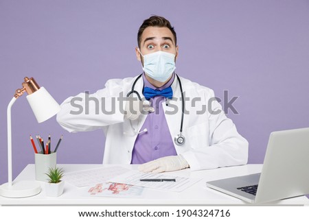 Shocked male doctor in white medical suit sit at desk work in clinic office in sterile mask coronavirus covid-19 and gloves point index finger on himself isolated on violet background studio portrait