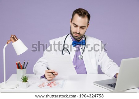 Male smart doctor man wearing white medical gown suit sits at desk work on computer in clinic office writing down reading documents papers internet isolated on violet purple background studio portrait