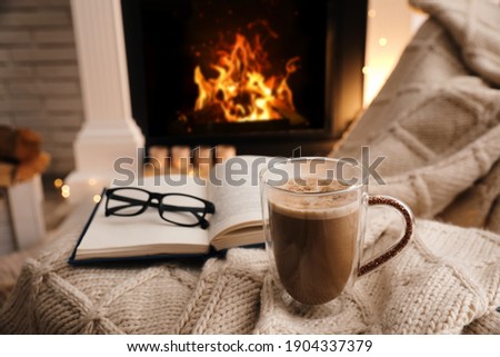 Cup of coffee, book and glasses near fireplace indoors, space for text. Cozy atmosphere