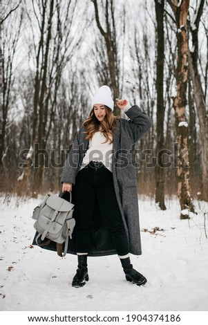 a blonde curly-haired girl dressed in a long dark black gray coat and a white hat walks along an alley in a snow-covered forest park in winter and holds a backpack in her hands