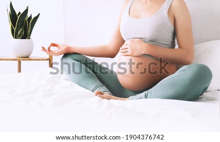Pregnant woman in lotus pose doing meditation or breathing exercises for healthy pregnancy and preparing body for childbirth. Young expectant mother practicing yoga at home. Royalty-Free Stock Photo #1904376742