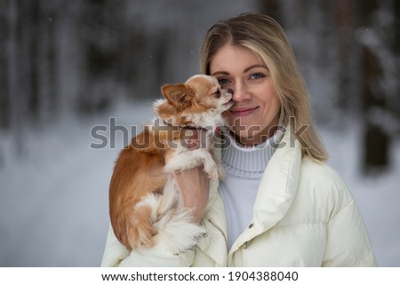 Blonde young female holding ginger and white chihuahua in her hands. Snow and trees on the background.