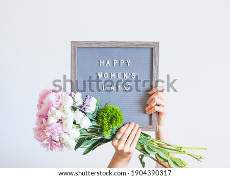 Young woman holding pink bouquet and letter board saying happy womens day on gray wall background