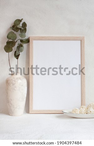 Blank sheet in a photo frame on the table. Vase with eucalyptus leaves, geometric balls on white plate. Mock up in a trendy style. Vertical template for minimal design.