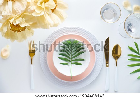 Effortless golden birthday dinner table decor. Pale yellow peony flowers and Late Spring, Summer flat lay. White dinner table, white and gold utensils, decorated with peony flowers and palm leaves.