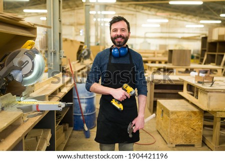 Smiling hispanic man doing carpentry work in a woodshop. Happy carpenter working with a power drill and a hammer Royalty-Free Stock Photo #1904421196