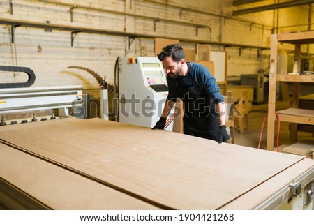 Male carpenter working with a plywood sheet in a big workshop. Handsome worker getting ready to cut a wood panel in a woodshop Royalty-Free Stock Photo #1904421268