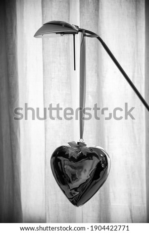 A heart-shaped helium balloon hangs on a room lamp. curtain in the background. decoration. black and white image.