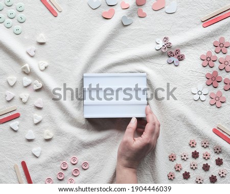 Hand hold lightbox with copy-space, text place. Flat lay with light box on ivory, off white textile. Hearts, candy, buttons, various decor items arranged on cotton tablecloth.