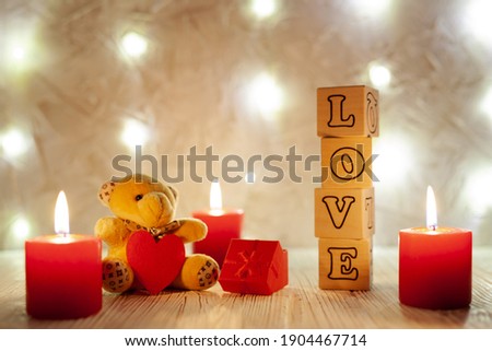 teddy bear, three burning red candles, wooden cubes with letters lined with the word love in the background with a burning garland