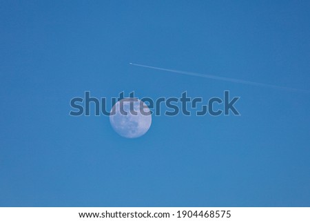 Moon in early evening with view of contrails of a plane flying over it. Moon on a clear blue sky. 