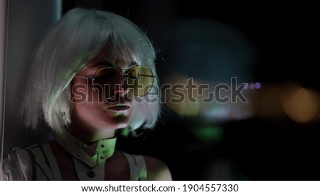 Close-up portrait of a beautiful blonde woman in stylish glasses sitting on the window sill against a dark background, thinking.