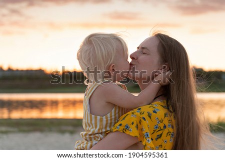 Young mother holding little daughter and gently kisses her. Portrait in sunset on river background.