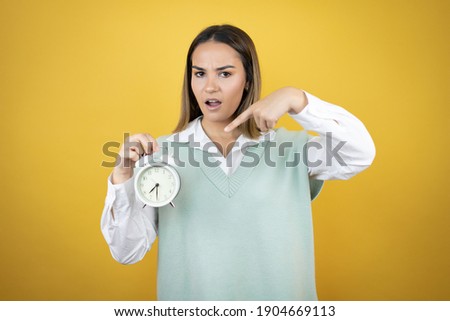 Pretty young woman standing over yellow background serious holding and pointing a clock