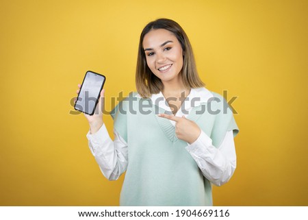 Pretty young woman standing over yellow background holding smartphone showing screen smiling happy pointing with hand and finger