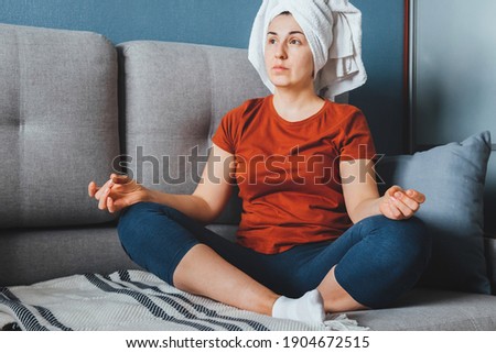 Girl with face mask and bath towel sitting on sofa and meditating in yoga pose yoga at home, quarantine, lockdown, meditation, mental health, wellness,self-care Film Grain