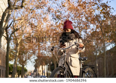Young African-American woman on her bicycle using her smartphone. She is wearing winter clothes, and is in a city with autumn weather.