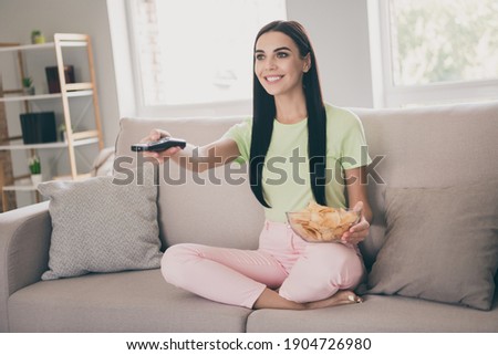 Photo of charming lady sit sofa crossed legs hold remote control bowl sliced potato white smile wear green t-shirt pink pants indoors