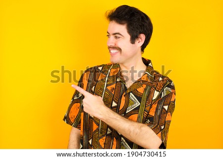 Happy cheerful smiling Young caucasian man wearing generic pattern printed shirt against yellow wall looking and pointing aside with hand. Copy space and advertisement concept.