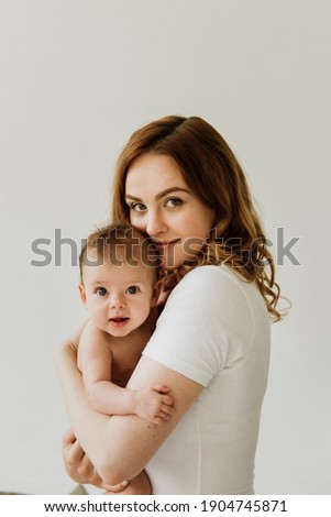 Beautiful mom hugs and kisses the newborn in the studio on a white background. A young mother in a white bodysuit hugs and kisses her newborn daughter in the studio.