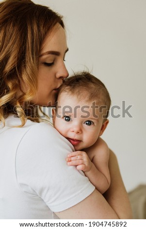 Beautiful mom hugs and kisses the newborn in the studio on a white background. A young mother in a white bodysuit hugs and kisses her newborn daughter in the studio. Royalty-Free Stock Photo #1904745892
