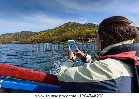 Pretty young woman taking photo on smartphone sailing lake Baikal on boat in autumn