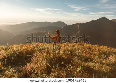 Carefree happy woman enjoying nature on grass meadow on top of mountain with sunrise, taking picture on her phone