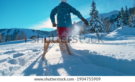 LOW ANGLE, CLOSE UP: Unrecognizable young woman drags a sled through deep snow on a sunny day. Excited female tourist enjoys her carefree winter vacation by running into fresh snow with her sleigh.