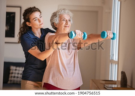 Old woman training with physiotherapist using dumbbells at home. Therapist assisting senior woman with exercises in nursing home. Elderly patient using dumbbells with outstretched arms. Royalty-Free Stock Photo #1904756515